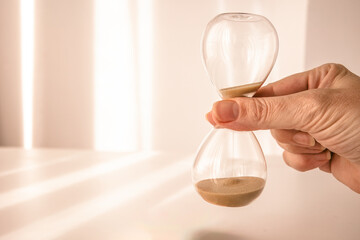 woman's hand holding time, depicted on an elegant glass hourglass with morning light background - obrazy, fototapety, plakaty