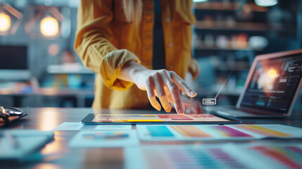A person is interacting with a futuristic holographic user interface spread over a desk, with a tablet, design palettes, and a laptop in a modern workspace.