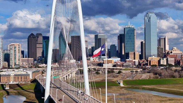 Texas Flag waving in the wind with Margaret Hunt Hill bridge and Dallas skyline in the background.