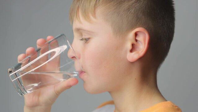 Cute boy drinks clean water from a glass on a gray background. portrait of a child with a glas