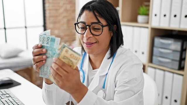 Smiling hispanic woman in a white lab coat holding brazilian real currency in a clinic office
