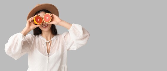 Beautiful young woman in stylish white dress and hat holding grapefruit on grey background with space for text