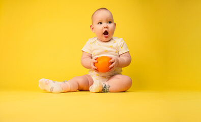 Beautiful baby girl holds orange sitting in studio on yellow background.