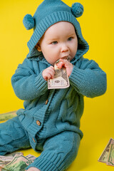 Cute kid chewing dollars on a yellow background. Baby in a blue knitted suit with money on a yellow background.