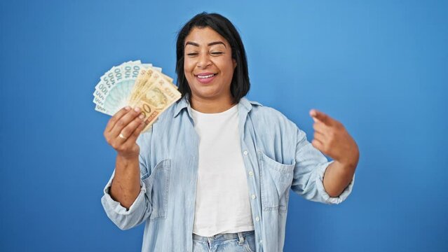 Smiling middle-aged hispanic woman holding brazilian reais against a blue background.