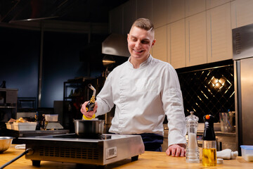 smiling chef stands in professional kitchen near the table about to set fire to alcohol in saucepans
