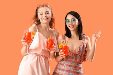Beautiful young women with glasses of tasty aperol spritz cocktail on orange background