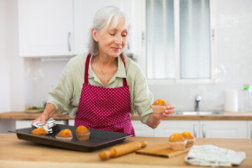 Positive elderly woman in apron standing in home kitchen holding baking tray with fresh muffins