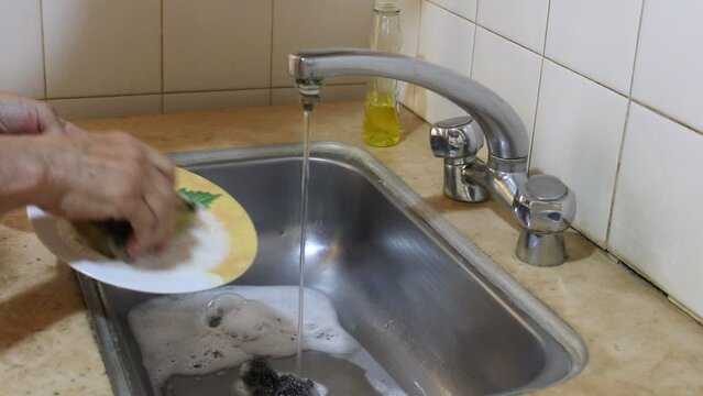 La mano del hombre lava los platos sucios del d&iacute;a anterior en el fregadero de casa, bajo el grifo de agua corriente mostrando c&oacute;mo la espuma deja el plato limpio, v&iacute;deos originales de la rutina diaria