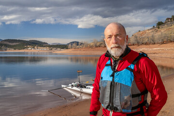 environmental portrait of a senior man wearing drysuit and life jacket with a rowing shell on lake...