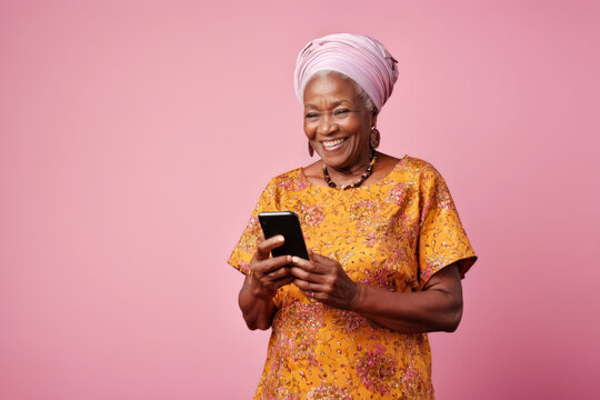 Portrait of smiling happy stylish African senior woman with turbant using mobile phone on pink background, studio shot with copy space for text