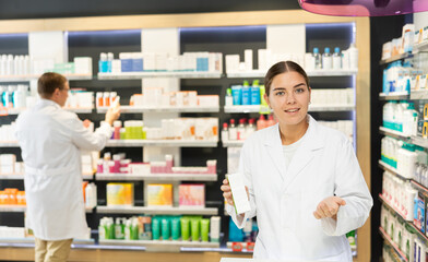 Polite young female pharmacist demonstrating preparation in box in chemist's shop with large assortment