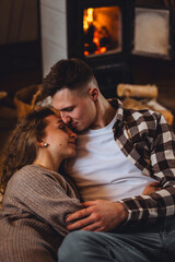 Happy young couple spending lazy weekend at home, embracing, kissing, enjoying near fireplace in the countryside cottage house, feeling relaxed and calm. Cozy home atmosphere, simple domestic life