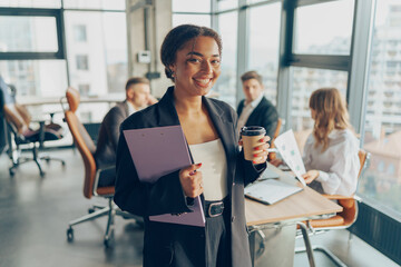 Smiling businesswoman in suit standing with coffee and clipboard at office on colleagues background