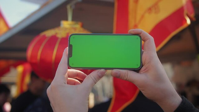 Young man holding phone chroma key screen on the street in front of chinese lignt close-up. Male with green screen on smart phone swiping . 