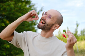 funny young charismatic man in beige t-shirt going to eat delicious ripe red cherries, green garden...