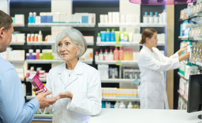 Polite mature female pharmacist consulting middle-aged man costumer about care product in box in...
