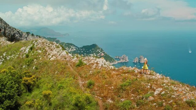 Lush greenery and rugged rocks with breathtaking view of the blue sea, with boats and rocky stacks of Faraglioni in the distance. Woman hiking in Capri, Italy.