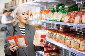 Focused positive elderly woman shopping in Asian food store, choosing traditional noodles for...
