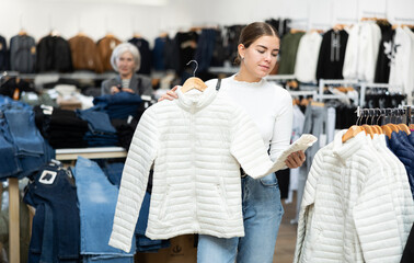 Pleased young girl choosing white jacket in clothing store with large assortment