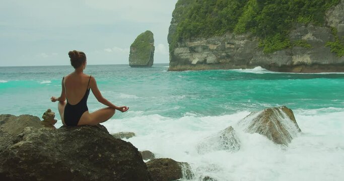 Serenity amidst ocean's roar. A woman meditates on a rocky beach, finding tranquility near turquoise ocean, waves crashing on cliffs.