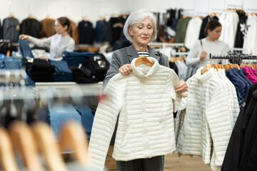 Pleased elderly woman choosing white winter down jacket in clothing shop with large assortment