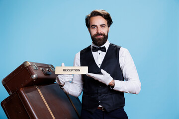 Hotel employee leading tourists in the right way using reception wall sign. Skilled professional bellboy posing on camera and holding front desk indicator, guiding guests and travellers.
