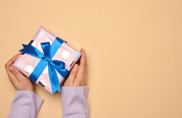 A woman's hand holds a gift box wrapped in a blue silk ribbon on a beige background, top view