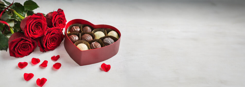 Caixa de chocolates em formato de cora&ccedil;&atilde;o, bouquet de rosas vermelhas, p&eacute;talas e fundo panor&acirc;mico branco, espa&ccedil;o para c&oacute;pia. Dia dos namorados.