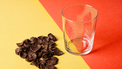 Chocolate corn flakes scattered on a yellow-red background, next to the empty transparent mug