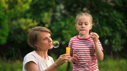 Loving mother with cheerful daughter blows bubbles in park among trees on vacation. Mother waits till preschooler girl blows bubbles in meadow on weekend. Mother and child spend time blowing bubbles