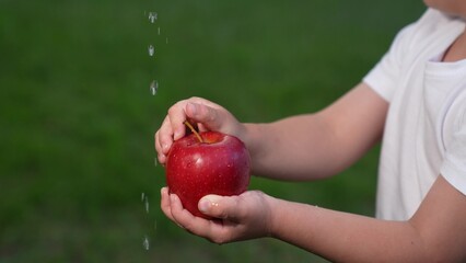 Male kid hands washing red ripe organic apple with water flow drops outdoor at summer park closeup. Boy child cleaning with aqua natural seasonal fruit edible plant enjoy healthy vitamin food garden