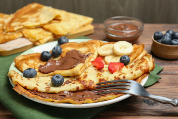 Tasty crepes with chocolate paste, banana and berries served on wooden table, closeup