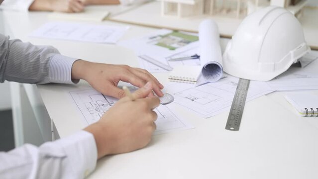 Close up of skilled architect engineer hand draws blueprint on meeting table with blueprint, architectural equipment, house model and safety helmet scatter around. Focus on hand. Immaculate.