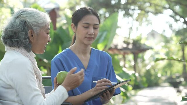 Compassionate Asian woman provides care to  elderly person in wheelchair outdoors. Engaging in physical therapy, happiness, encouraging positive environment for mature individuals with grey hair.