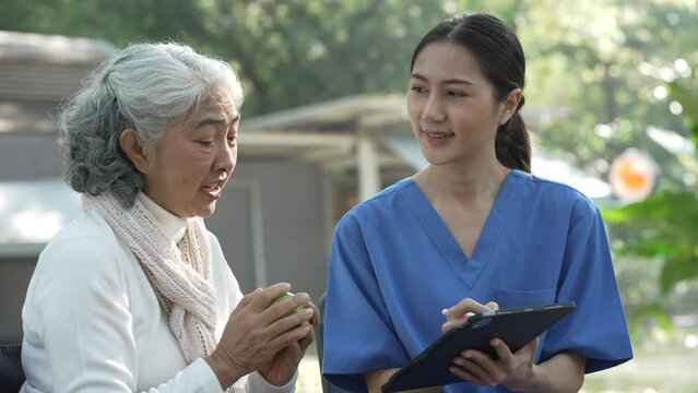 Compassionate Asian woman provides care to  elderly person in wheelchair outdoors. Engaging in physical therapy, happiness, encouraging positive environment for mature individuals with grey hair.