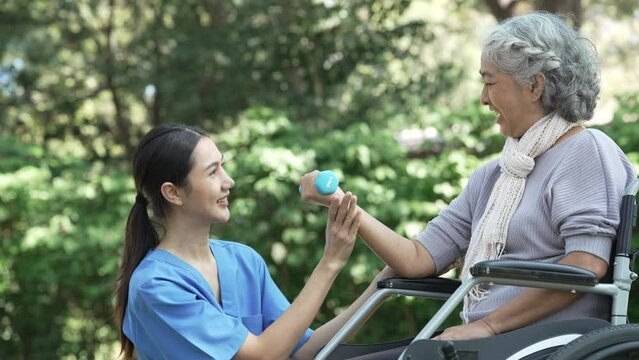 Compassionate Asian woman provides care to  elderly person in wheelchair outdoors. Engaging in physical therapy, happiness, encouraging positive environment for mature individuals with grey hair.