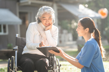 Compassionate Asian woman provides care to  elderly person in wheelchair outdoors. Engaging in...