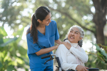 Compassionate Asian woman provides care to  elderly person in wheelchair outdoors. Engaging in...