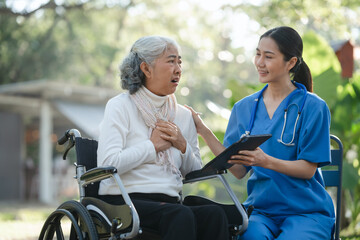 Fototapeta premium Compassionate Asian woman provides care to elderly person in wheelchair outdoors. Engaging in physical therapy, happiness, encouraging positive environment for mature individuals with grey hair.