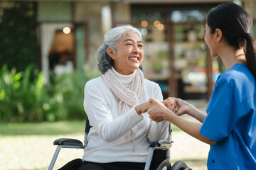 Compassionate Asian woman provides care to  elderly person in wheelchair outdoors. Engaging in...