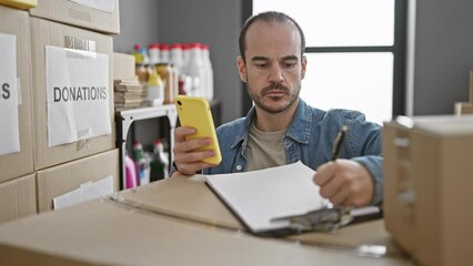 A focused bald hispanic man with a beard organizing donations in a room while using a smartphone. - Powered by Adobe