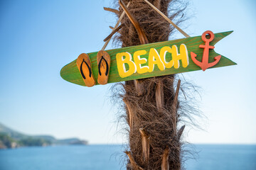 Green shaped sign with the word 'BEACH' pointing the way, tied to a palm tree with the ocean in the distance
