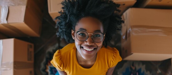 A woman with glasses and a yellow shirt sits among cardboard boxes, smiling happily. She is enjoying the event and having fun, sharing a moment captured in a photo caption - obrazy, fototapety, plakaty