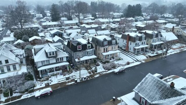Snowflakes over a residential street with row houses, bare trees, and parked cars. Aerial approach towards historic town housing during winter snow storm in USA.
