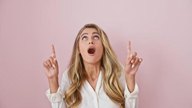 Young blonde woman, looking up in sheer amazement, pointing with finger over isolated pink background. standing in shirt, she oozes a joyful, confident, standing-surprise look