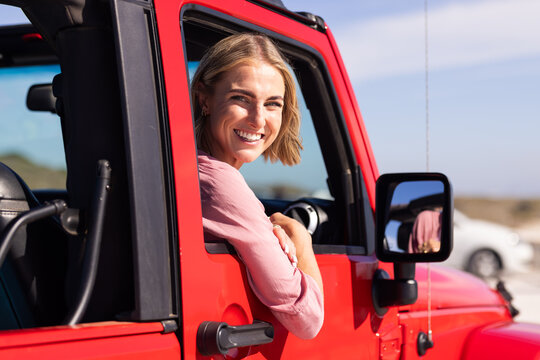 Young Caucasian woman smiles from the driver's seat of a red vehicle on a road trip