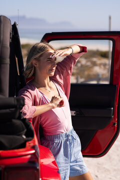 Young Caucasian woman shields her eyes from the sun near a red vehicle on a road trip