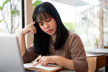 A serious and concentrated young Asian woman is reading a book in a cafe.