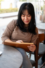 A positive Asian female is keeping her diary or listing her ideas in a book while sitting in a cafe.